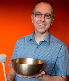 Ein lächelnder Mann hält eine Klangschale und einen Schlegel vor einer orangefarbenen Wand.