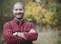 Man in a red plaid shirt smiling with crossed arms, standing outside with autumn trees behind.