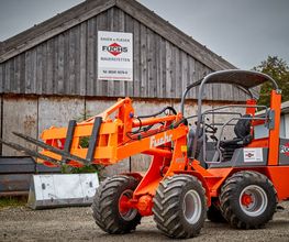 Orange Radlader vor Holzgebäude mit Fuchs-Logo und Kontaktdaten, grauer Himmel im Hintergrund.