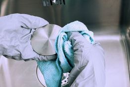 Zwei behandschuhte Hände reinigen ein rundes Metallsieb mit einem blauen Tuch über einem Spülbecken.