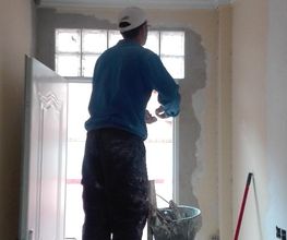 Persona pintando pared en escalera dentro de una habitación. Lleva gorra blanca y ropa de trabajo.