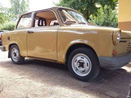 Beiger Trabant-Pkw parkt auf einer Einfahrt, umgeben von Bäumen und einem Haus im Hintergrund.