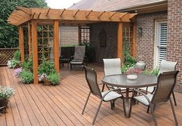 Wooden deck with pergola, patio table, chairs, potted plants, and brick wall background.
