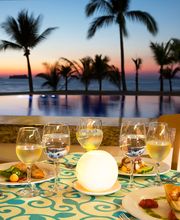 Cena junto al océano al atardecer, con mesa servida y copas de vino, palmeras de fondo.