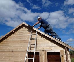Man wearing a hard hat working on a wooden roof under a blue sky with clouds.