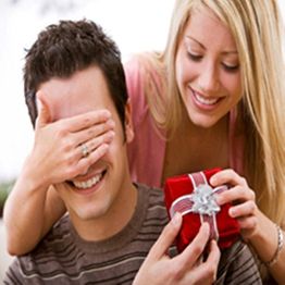 Woman surprises man with gift, covering his eyes while he smiles, holding a red present.