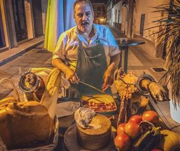 Hombre en delantal con mesas de embutidos, queso y verduras en una calle iluminada.