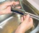 Hands fixing a metal kitchen faucet above a stainless steel sink.