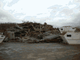 Ein Berg aus Holzresten und Trümmern auf einem Parkplatz unter bewölktem Himmel.