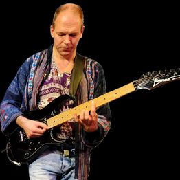 Person spielt E-Gitarre auf der Bühne, trägt buntes Hemd und Jeans, schwarzer Hintergrund.