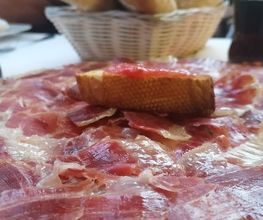 Plato de jamón ibérico con pan y bebidas en la mesa, cesta con pan al fondo.