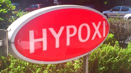 Rotes Schild mit der Aufschrift "HYPOXI" vor einer grünen Hecke und mehreren geparkten Autos.