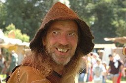 Mann mit braunem Hut lächelt draußen, im Hintergrund sind Bäume und unscharfe Personen.