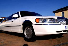Weiße Limousine auf einem gepflasterten Parkplatz vor einem Gebäude bei sonnigem Wetter.
