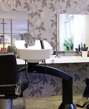 Friseursalon mit Waschbecken, Spiegel, zwei Stühlen und Pflanzen auf dem Tisch vor gemusterter Wand.