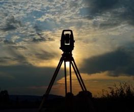 Vermessungsgerät auf Stativ bei Sonnenuntergang, vor bewölktem Himmel.