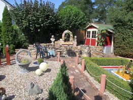 Ein bunter Garten mit Steinskulpturen, kleinen Büschen und einem roten Gartenhaus im Hintergrund.