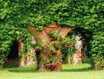 Bewachsene Backsteinmauer mit Arkaden und roten Blumen, umgeben von grünem Laub.