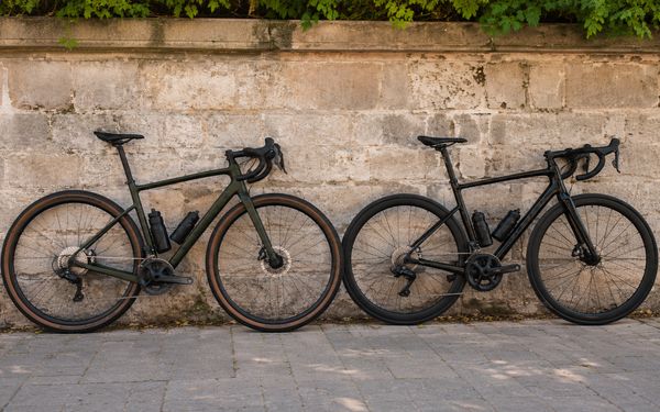 Gravel Bike und Rennrad lehnen nebeneinander an einer alten Steinmauer auf einem gepflasterten Gehweg.