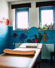 Behandlungsraum mit maritimer Wandmalerei, Liege, zwei Fenster, Bücher und Spielzeug.