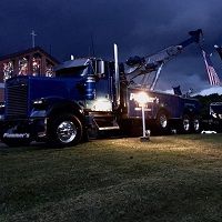 Blue tow truck at night with lit buildings and American flag under a dark, cloudy sky.