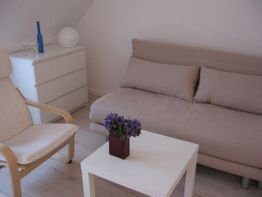 Wohnzimmer mit beigem Sofa, weißem Tisch, Sessel, Kommode und violetten Blumen in einer Vase.
