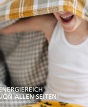 Kind lacht unter einer bunt karierten Decke. Text: "Energierich von allen Seiten!"