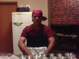 Homem de avental vermelho e touca, sentado com potes de vidro em uma cozinha com churrasqueira.