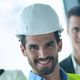 Smiling man in a hard hat and vest, standing indoors with a blurred background.