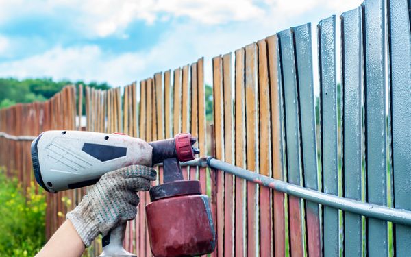 Ein Mann lackiert einen Zaun mit einem Farbsprühgerät. Lackierung des Zauns zum Schutz vor Korrosion.