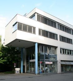 Modernes weißes Gebäude mit großen Fenstern und blauen Säulen, Geschäfte im Erdgeschoss.