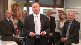 Eine Gruppe von sechs Personen in Business-Kleidung posiert vor Werbebannern in einem Büro.