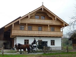 Zweistöckiges Holzhaus, davor Pferdekutsche mit zwei Pferden und Kutscher. Rustikale Umgebung.