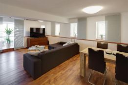 Wohnzimmer mit Holzboden, braunem Sofa, Esstisch, Stühlen, Fernseher und Pflanzen am Fenster.
