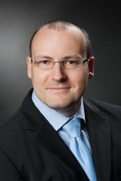 Ein Mann mit Brille trägt Anzug und Krawatte, vor neutralem Hintergrund.