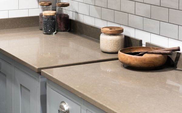Encimera de cocina de cuarzo en tono beige, con superficie lisa y pulida, acompañada de tarros de vidrio con alimentos y un bol de madera sobre muebles blancos y salpicadero de azulejos.