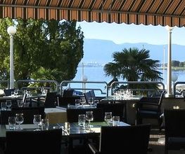 Restaurantterrasse am See mit gedeckten Tischen, Sonnenschirm, Bäumen und Bergblick im Hintergrund.