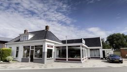 Wit gebouw met winkelruiten aan een straat, auto geparkeerd rechts, met bewolkte lucht erboven.