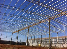 Estrutura metálica de um galpão em construção, céu azul ao fundo.