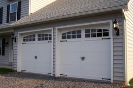 White double garage doors with decorative windows on a white house, exterior light fixtures mounted.