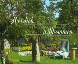 Gemütlicher Garten mit Liegestuhl, Tisch und Pflanzen; Text: "Herzlich willkommen".