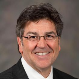 Man in a suit smiling, wearing glasses against a neutral background.