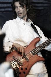 Ein Mann spielt Bassgitarre in einem weißen Hemd. Er hat lange, dunkle Haare.