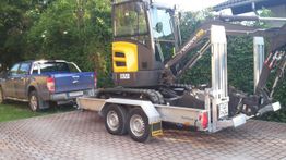 Blauer Pickup zieht gelben Mini-Bagger auf einem Anhänger vor grüner Hecke.