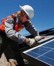 Persona instalando panel solar con casco y chaleco, cielo despejado de fondo.