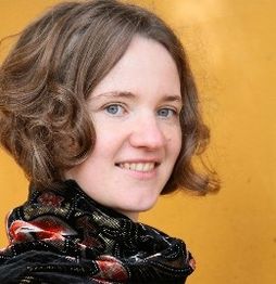 Eine lächelnde Frau mit kurzen Haaren vor einem orangefarbenen Hintergrund.