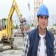 Smiling construction worker in blue hard hat, with excavator in the background.