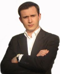 Mann im Anzug schaut ernst mit verschränkten Armen vor neutralem Hintergrund.