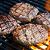 Grilled steak patties on a barbecue with char marks and flames beneath.