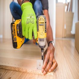 A person in green gloves using a yellow power drill on wooden boards indoors.